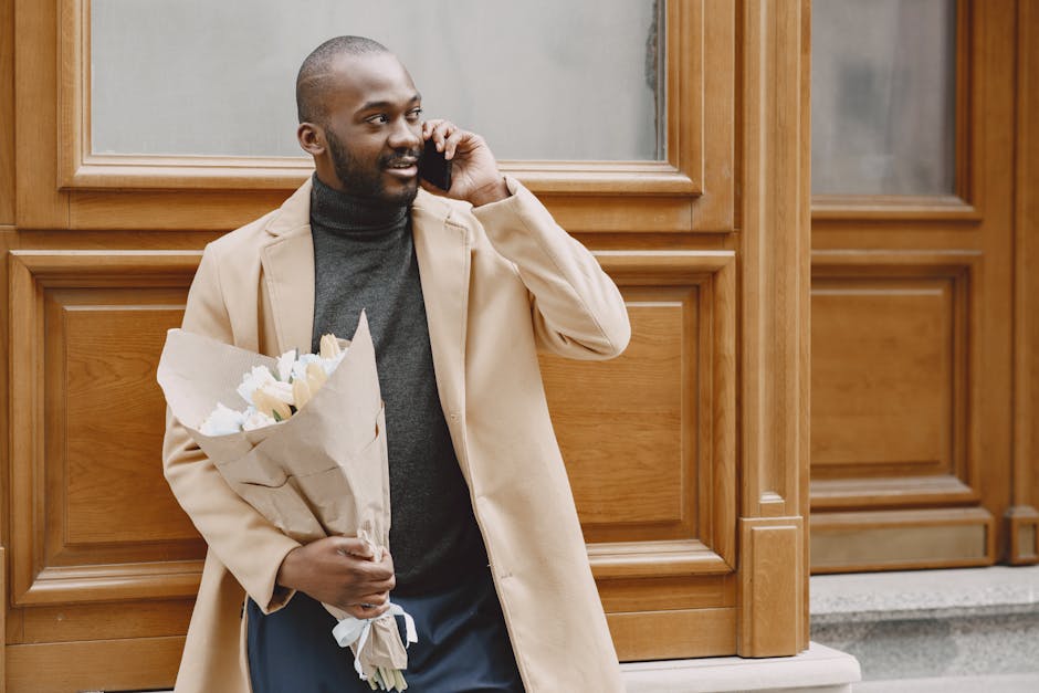 A man with a dark complexion, short hair, and a beard, dressed in a beige coat over a dark turtleneck, is standing outdoors against a wooden-paneled wall. He is holding a bouquet of fresh white flower