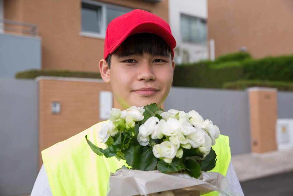 A young male flower delivery courier wearing a red cap and high-visibility yellow vest, smiling and holding a bouquet of white roses wrapped in white paper, with green leaves visible among the flowers