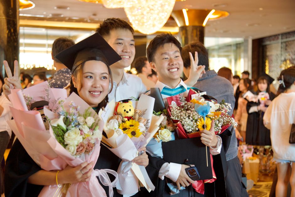 Three graduates at a celebration event, holding vibrant floral bouquets including pink and white roses, sunflowers, and baby's breath, wrapped in pastel pink and white paper with ribbons. One of them,