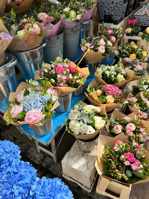 A display of fresh floral bouquets arranged in metal buckets, paper wraps, and vases on a table at a flower market. The arrangements feature a variety of flowers, including pink roses, white and pale 