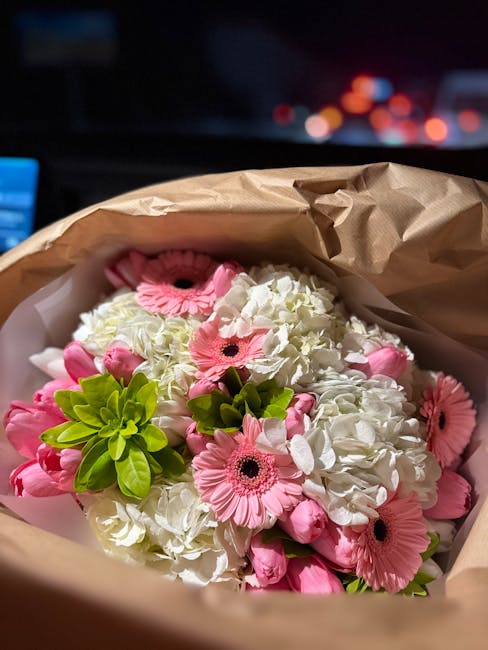A bouquet of fresh pink gerbera daisies and white hydrangeas, accented with light green foliage, arranged in a compact floral display. The flowers are enclosed in white tissue paper and wrapped with b