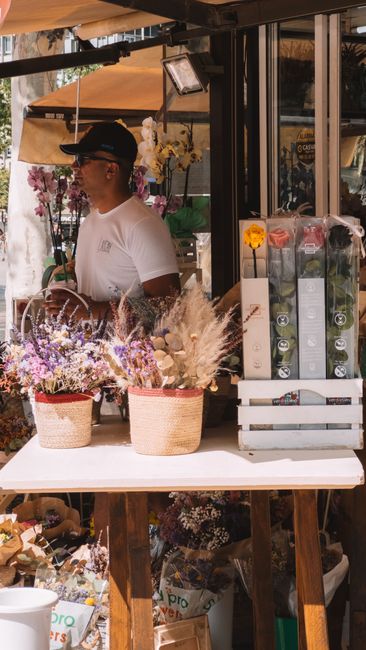A florist at a flower stall on Uxbridge High Street arranging a bouquet of fresh flowers. The display features a variety of blooms including purple lavender, white and pink flowers, and dried pampas g